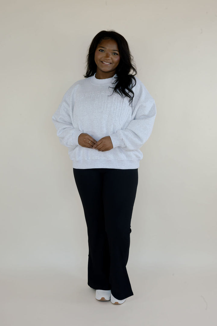 Full-length view of woman wearing heather gray Simply For Good crewneck sweatshirt with black flare pants and white shoes