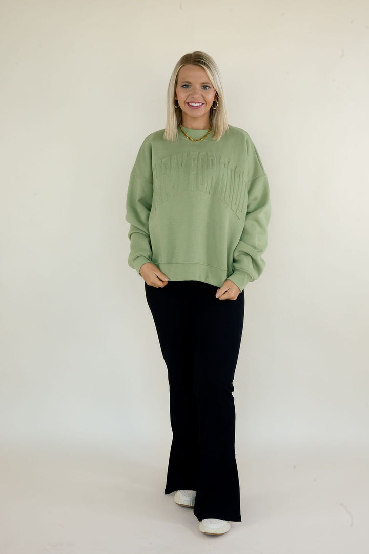 Woman wearing sage green Simply For Good crewneck sweatshirt with tonal front lettering and black flare pants
