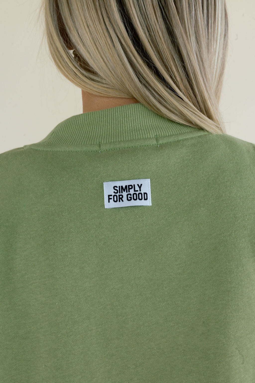 Close-up of sage green sweatshirt showing small Simply For Good branding at the back neckline