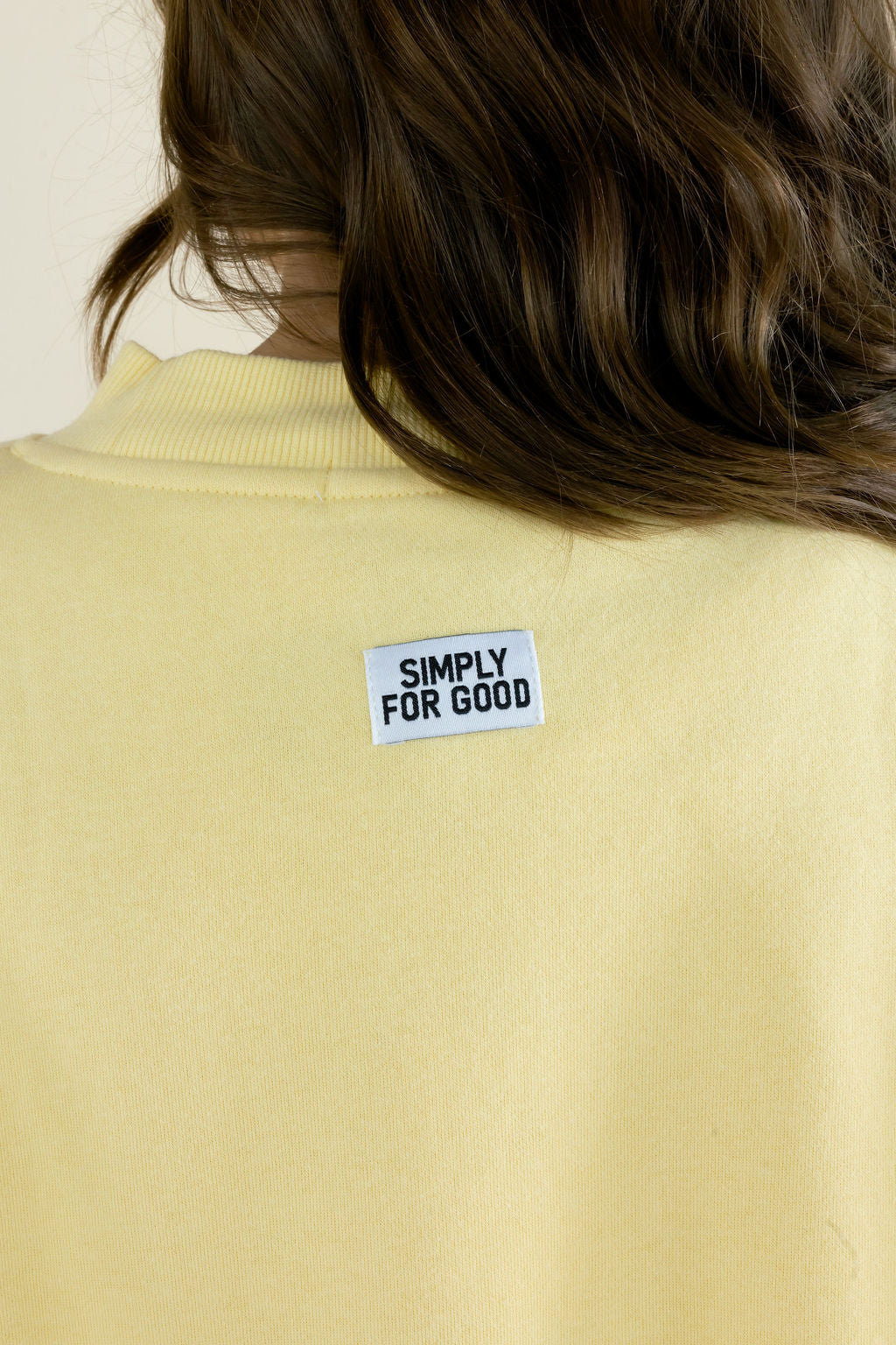 Close-up of butter yellow sweatshirt showing small Simply For Good branding centered below the back neckline