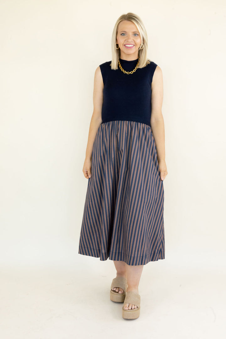 Woman modeling navy sleeveless midi dress with solid knit bodice and flowy navy and brown striped skirt styled with neutral platform sandals