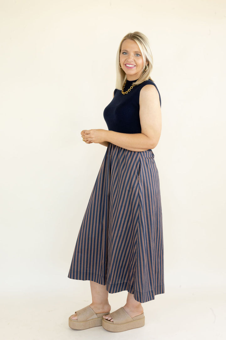 Side view of woman wearing navy sleeveless midi dress with solid knit bodice, striped gathered skirt, and mid-calf length