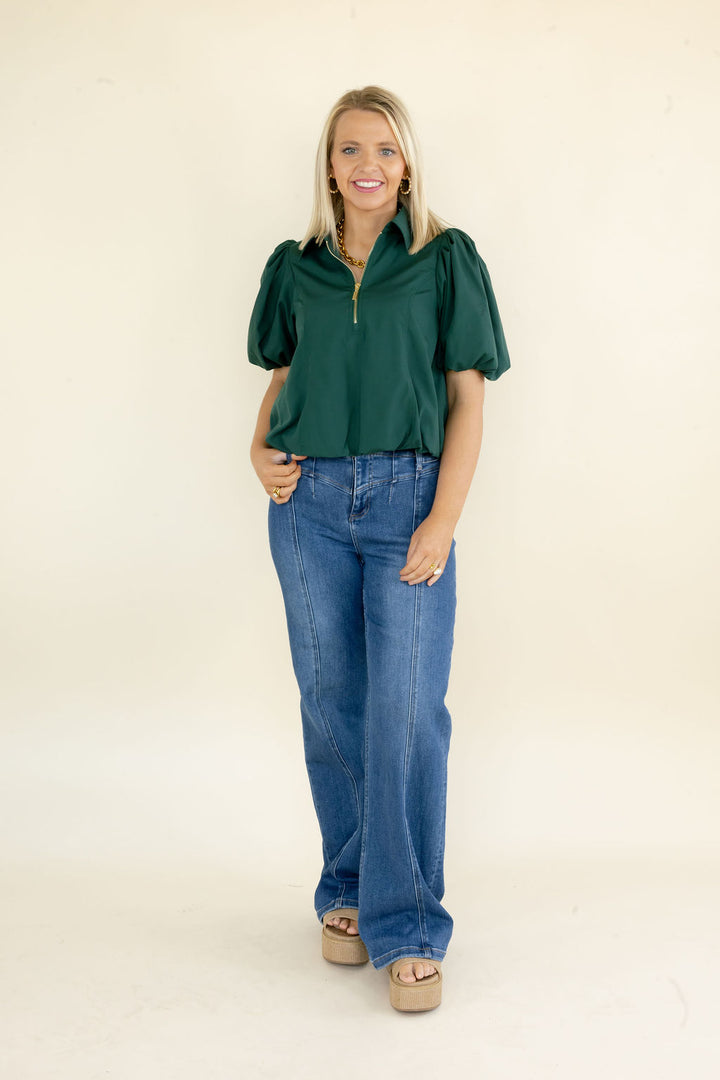 A woman modeling a dark green short-sleeve blouse with puff sleeves and a half-zip neckline, paired with blue jeans and beige platform sandals, against a plain cream background.
