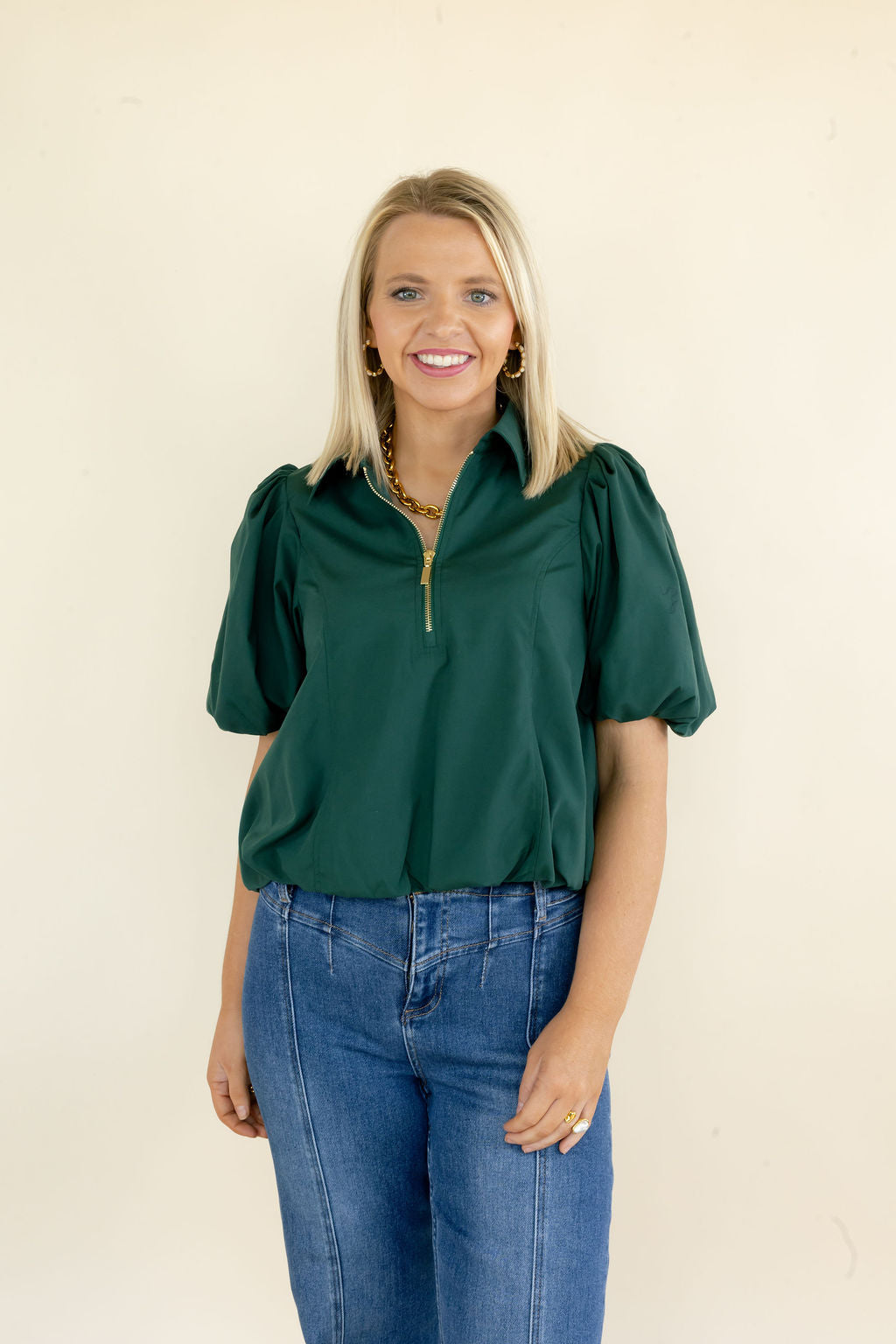 A smiling blonde woman wearing a dark green short-sleeve blouse with puff sleeves and a gold zipper at the collar, paired with blue jeans, standing against a plain off-white background.