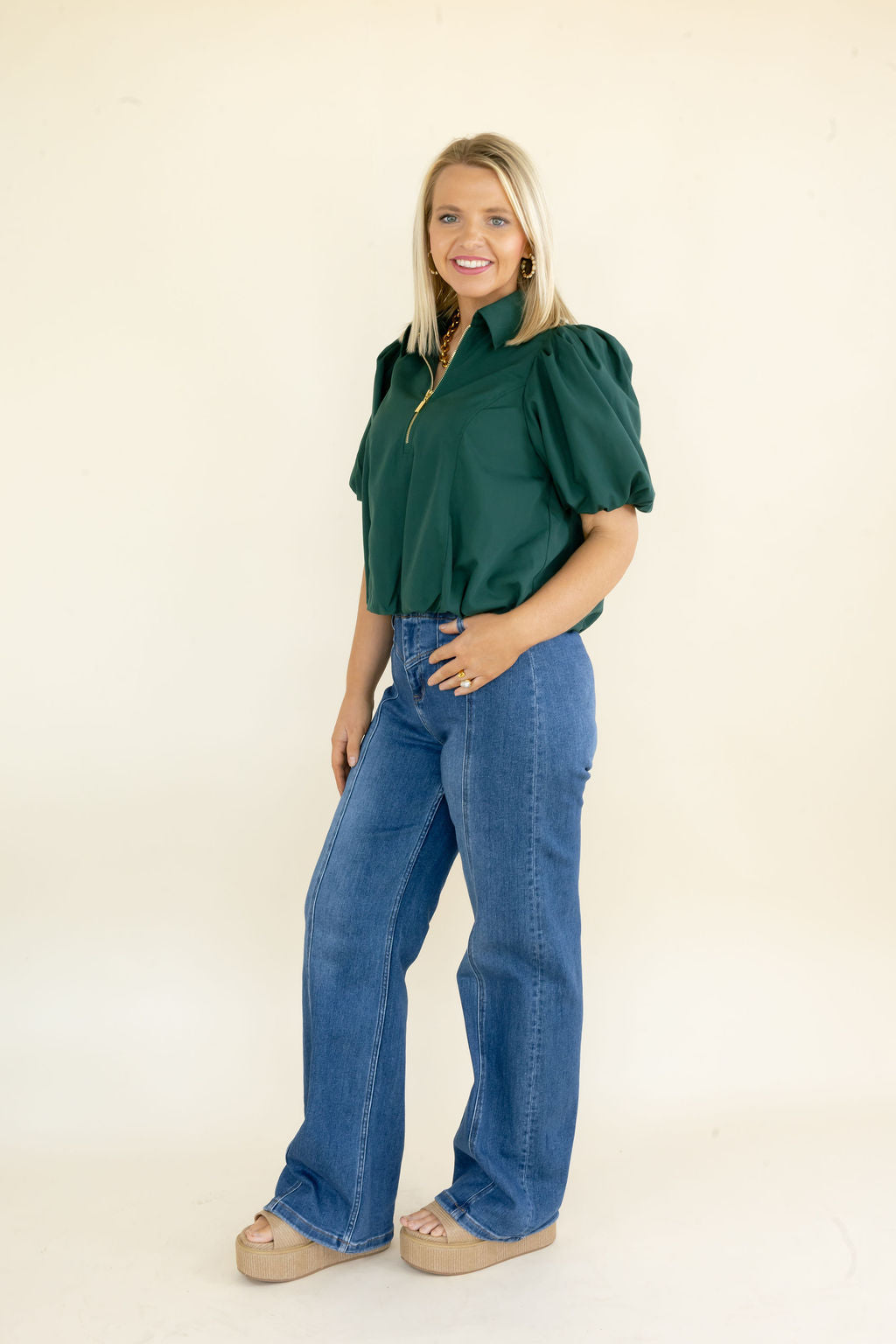 A woman modeling a dark green puff-sleeve blouse with a gold zipper, paired with wide-leg blue jeans and beige platform sandals, standing against a plain cream background.