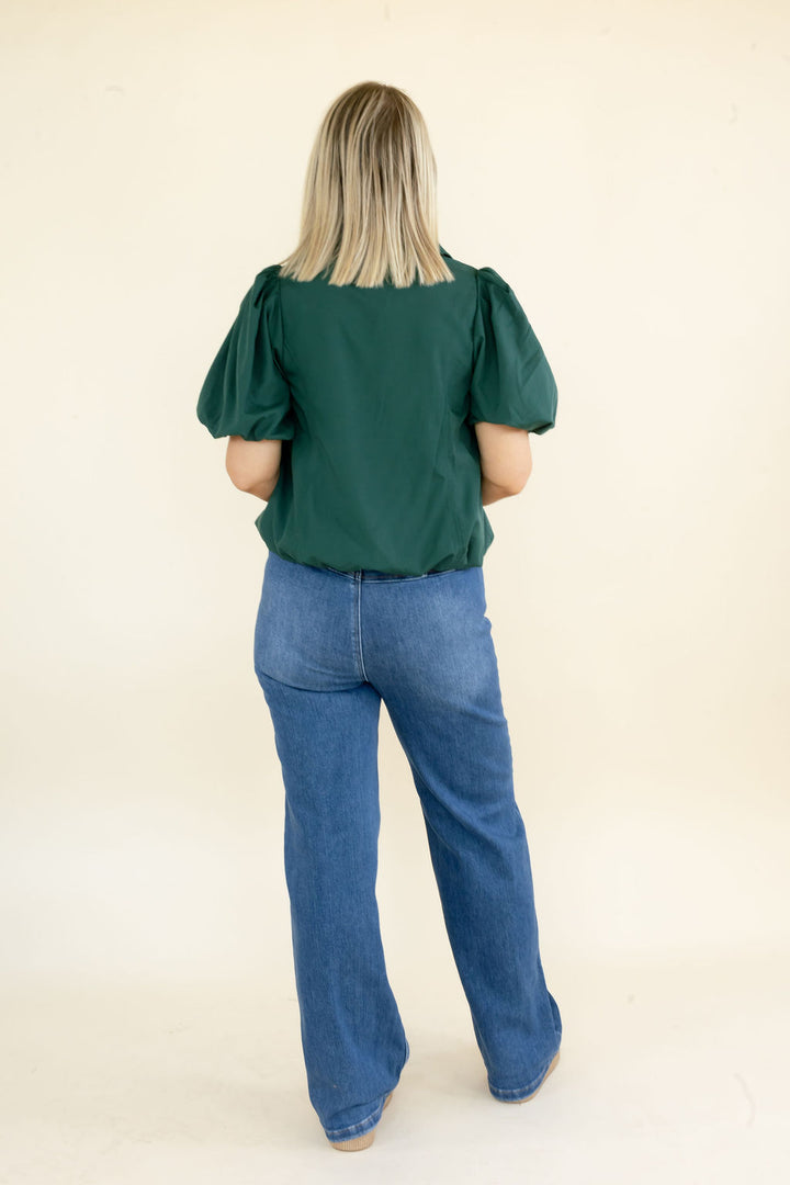 A woman viewed from behind wearing a dark green puff-sleeve top and blue jeans, standing against a plain cream-colored studio background.