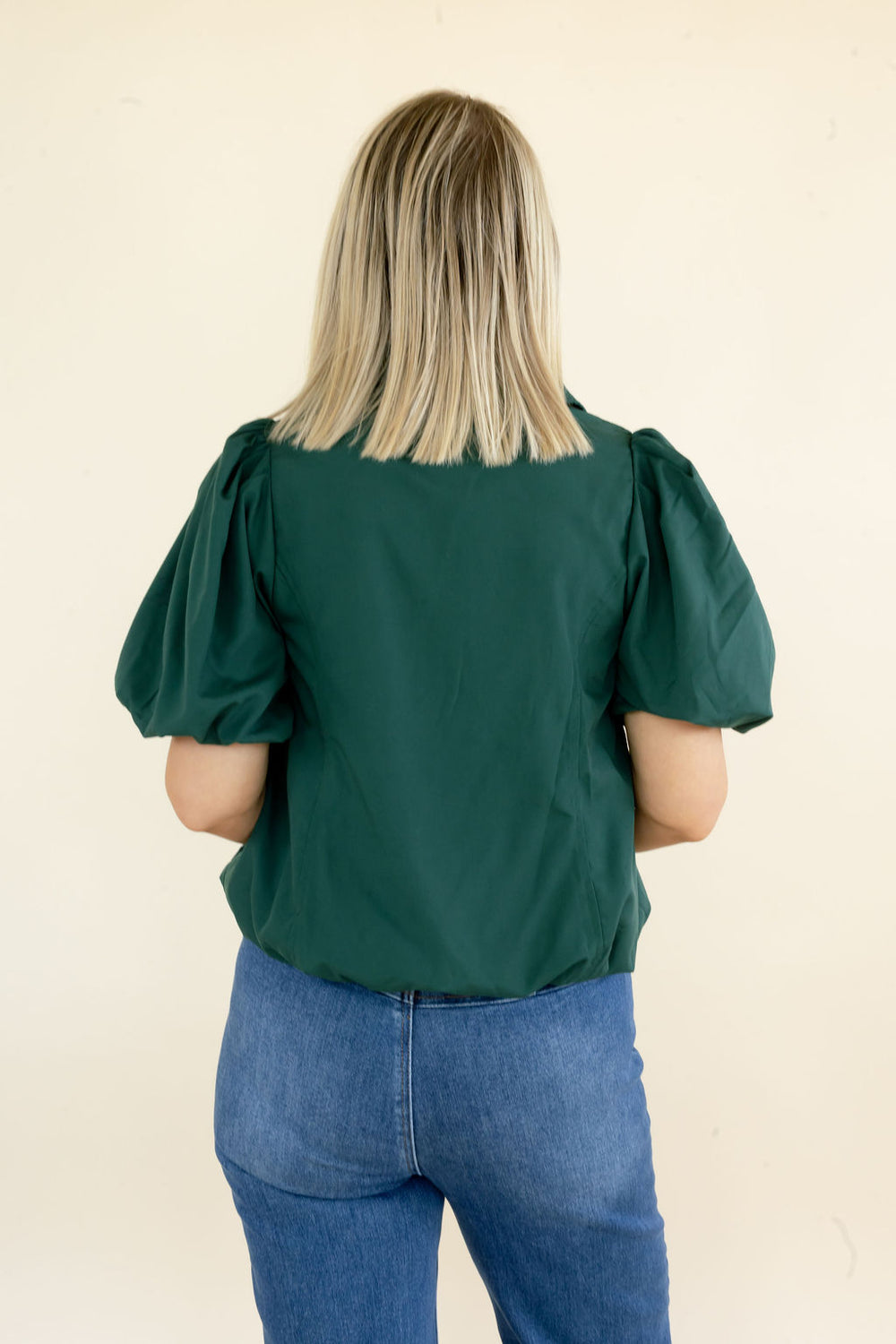 A woman viewed from behind wearing a dark green puff-sleeve blouse and blue jeans, standing against a plain cream-colored wall.