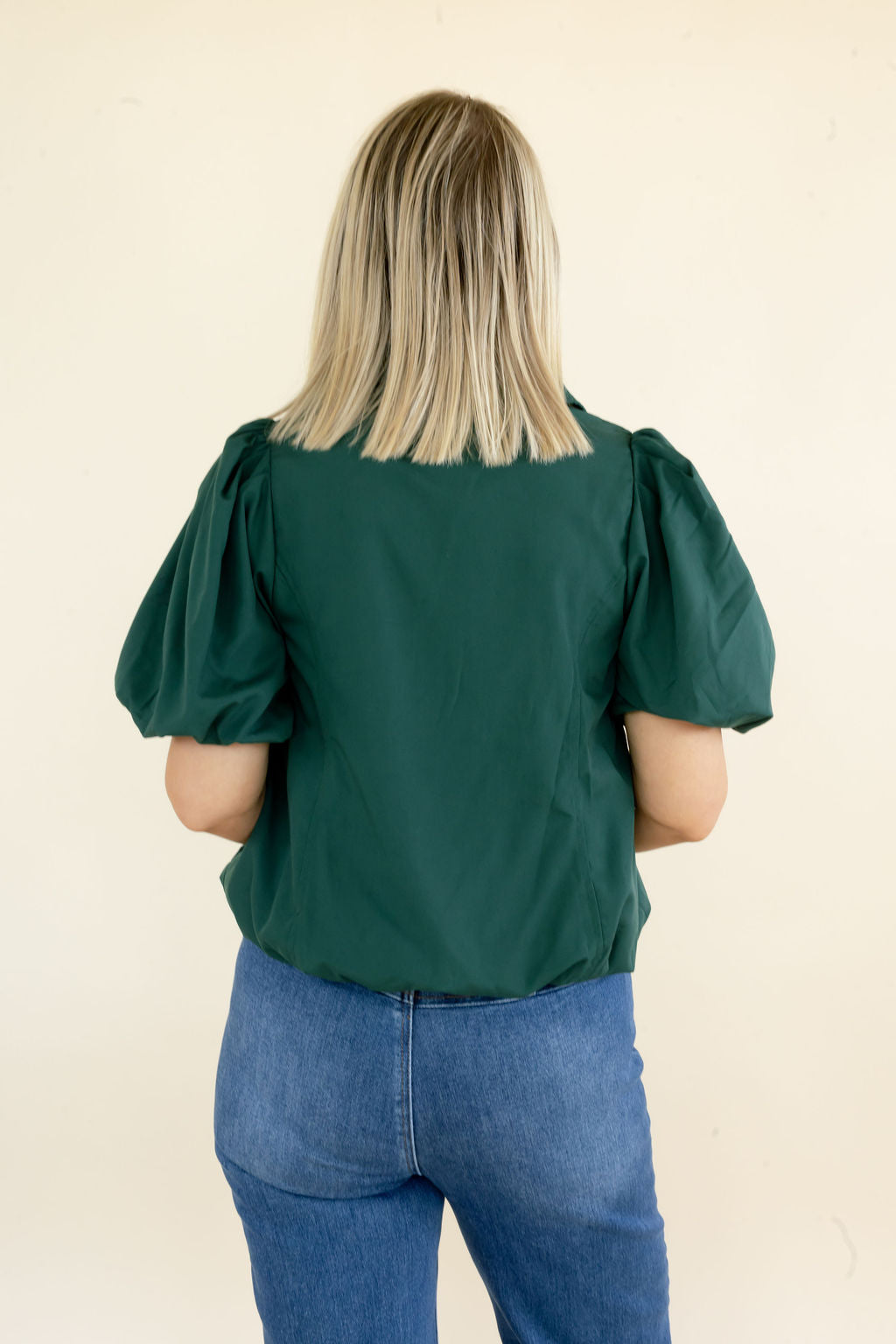 A woman viewed from behind wearing a dark green puff-sleeve blouse and blue jeans, standing against a plain cream-colored wall.