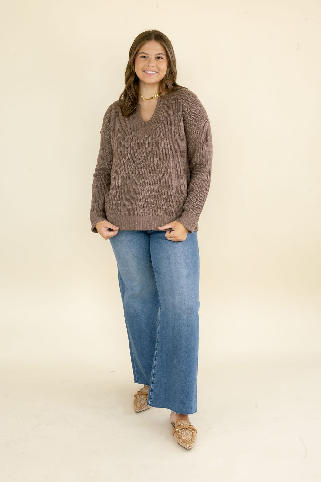 Woman modeling taupe brown V-neck knit sweater with long sleeves and relaxed fit styled with blue wide leg jeans and neutral flats