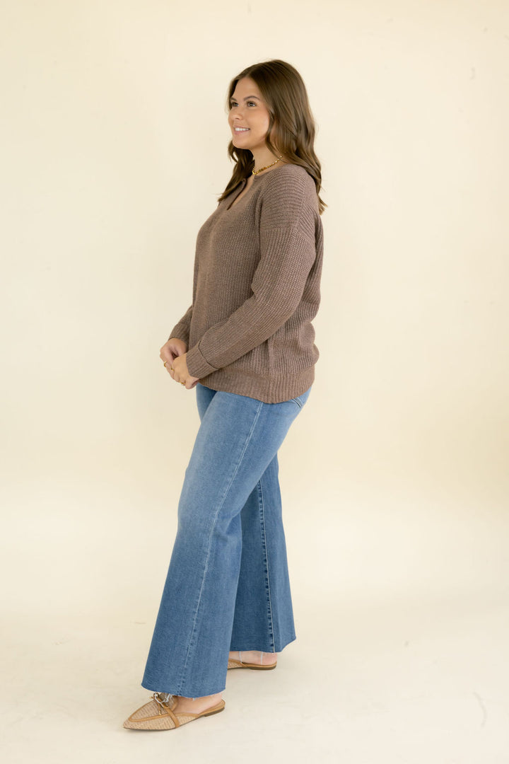 Side view of woman modeling taupe brown V-neck knit sweater with relaxed fit styled with blue wide leg jeans