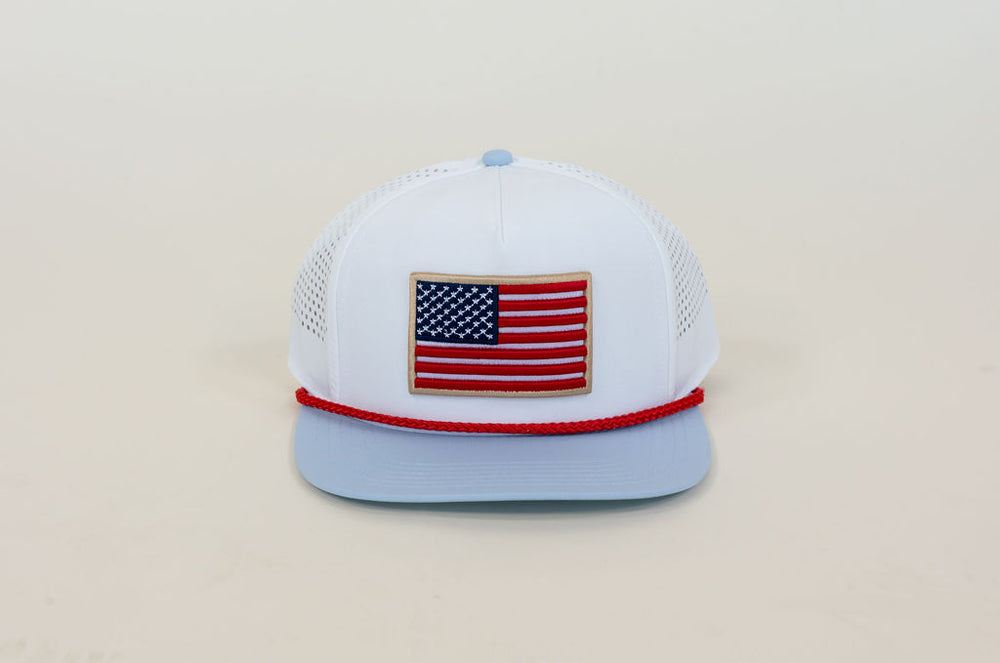 White performance trucker hat featuring embroidered American flag patch, light blue flat bill, red rope trim, and breathable mesh panels
