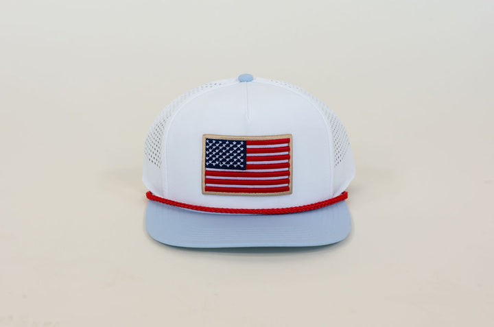 White performance trucker hat featuring embroidered American flag patch, light blue flat bill, red rope trim, and breathable mesh panels