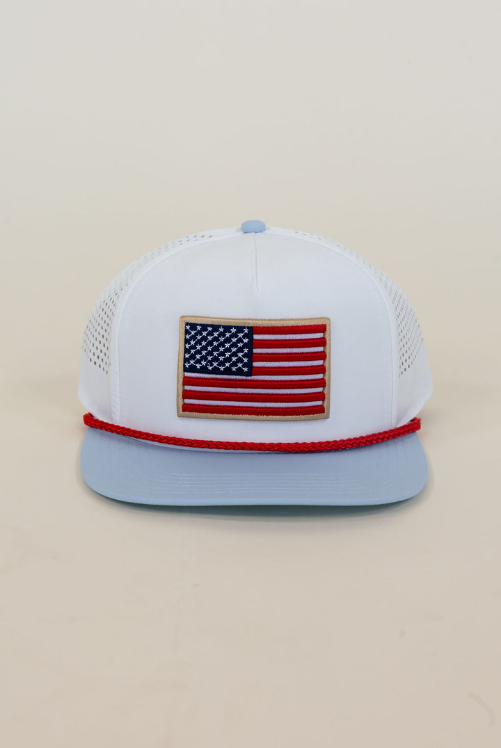 Close-up of white American flag trucker cap featuring red, white, and blue embroidered flag patch, red rope detail, light blue bill, and perforated mesh sides