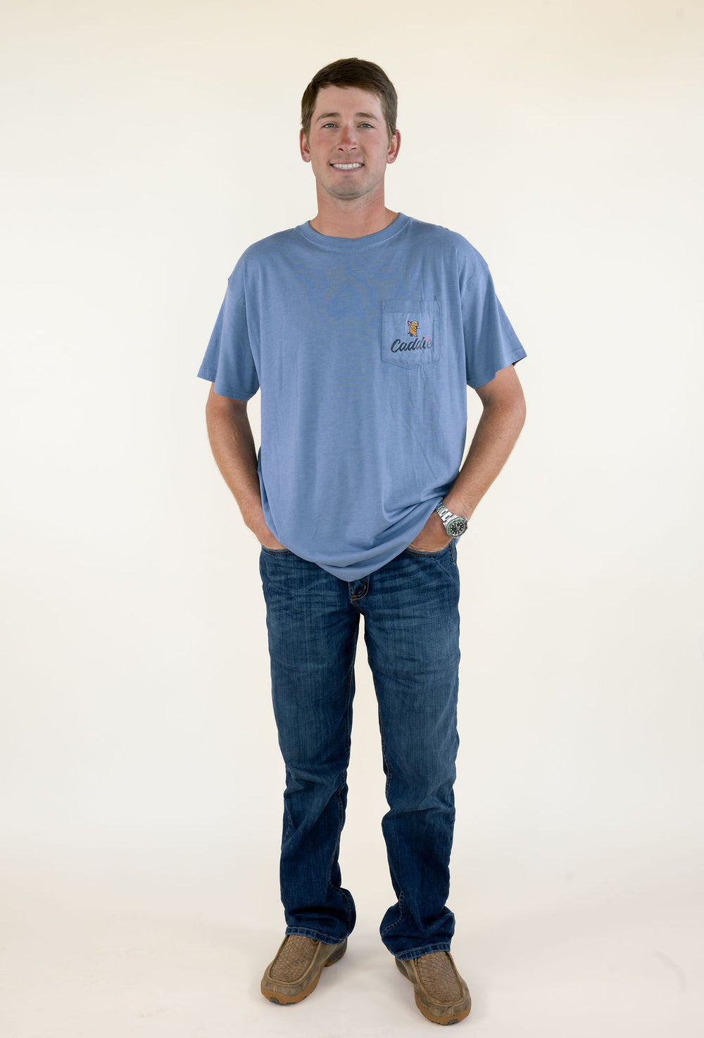 Full-length front view of man wearing blue Caddie short sleeve graphic T-shirt with jeans and brown boots