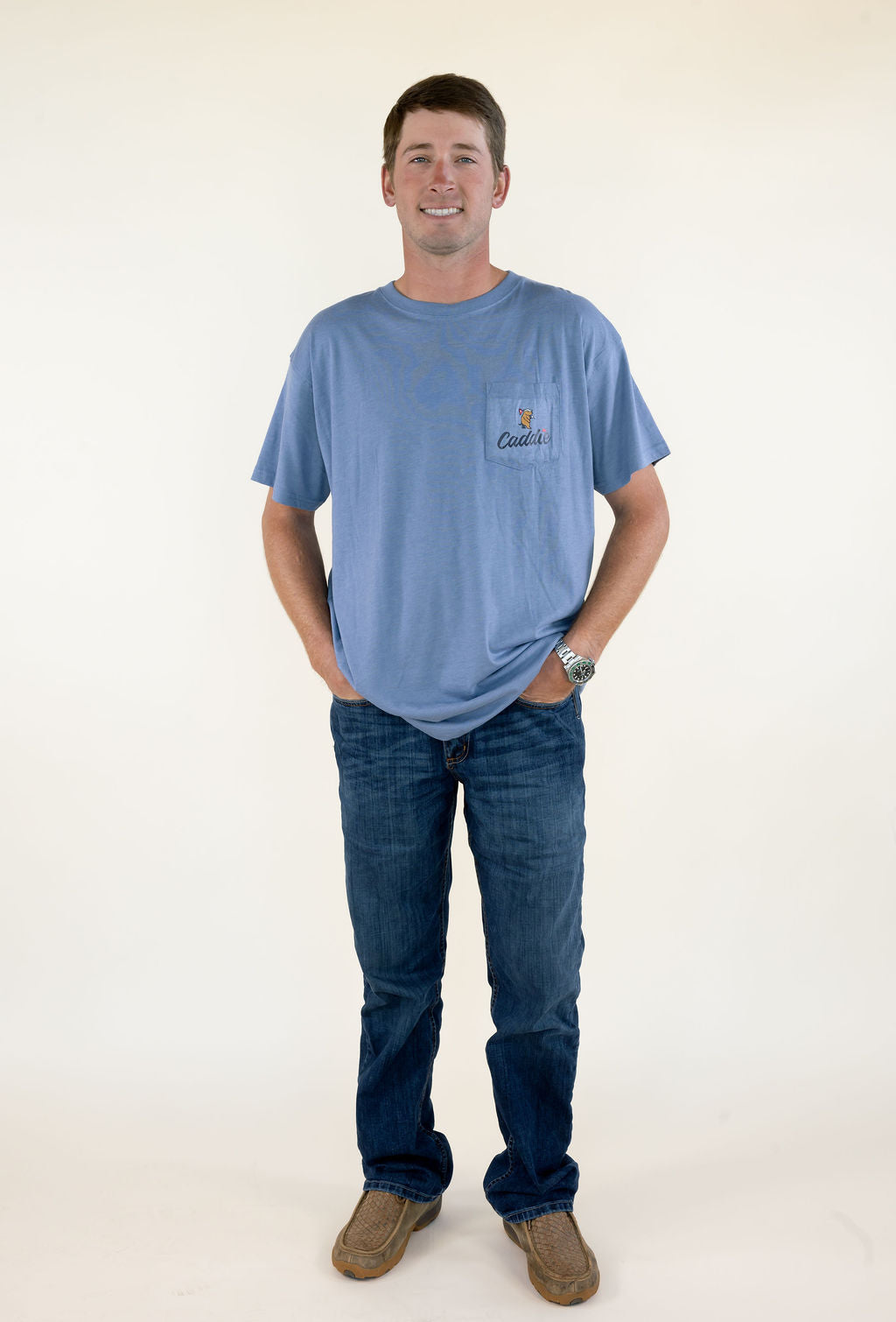 Full-length front view of man wearing blue Caddie short sleeve graphic T-shirt with jeans and brown boots