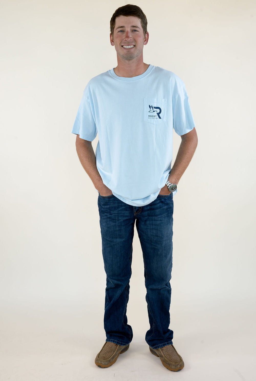 Full-length front view of man wearing light blue Roost Waterfowl Supply Co. short sleeve T-shirt with jeans and brown boots