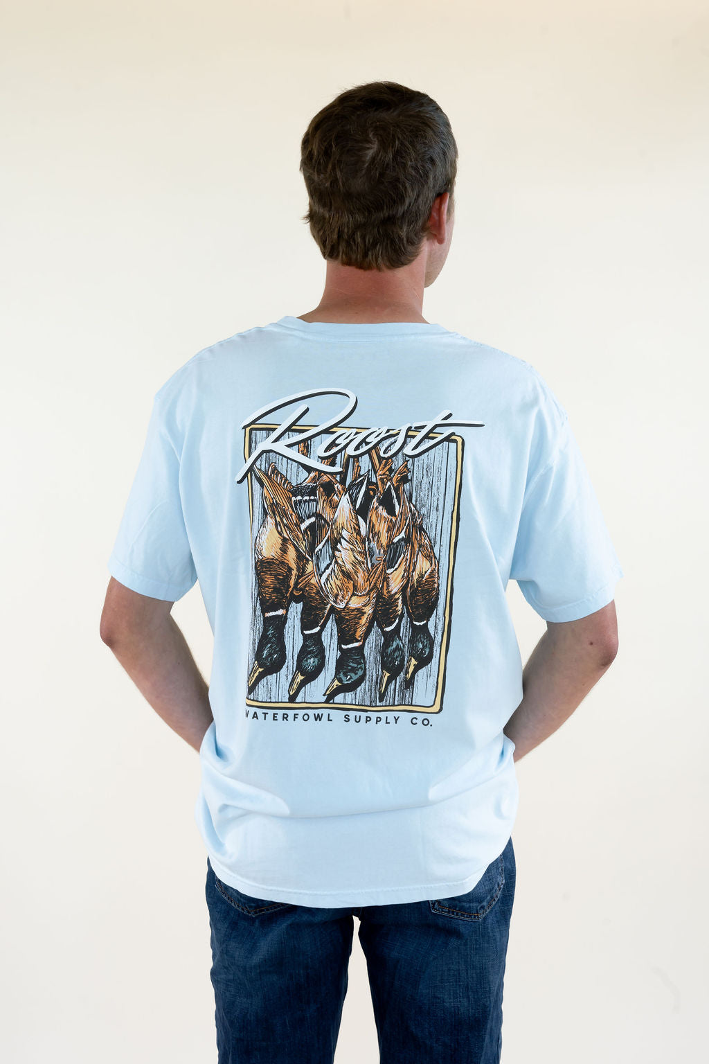 Man modeling light blue Roost Waterfowl Supply Co. graphic T-shirt featuring a large duck hunting back graphic, short sleeves, and relaxed fit