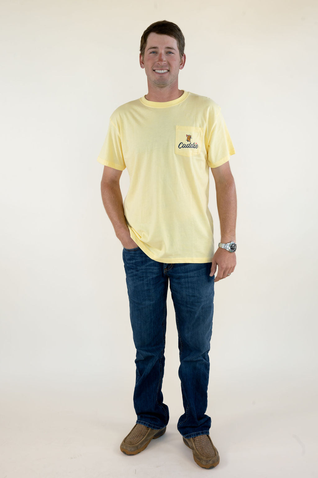 Full-length front view of man wearing pale yellow Caddie short sleeve golf T-shirt with blue jeans and brown boots