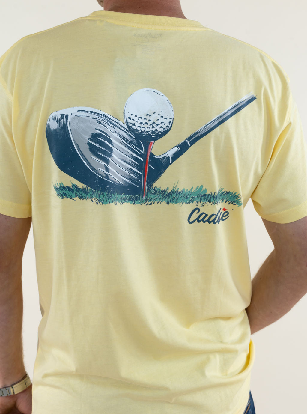 Close-up back view of pale yellow Caddie graphic T-shirt highlighting the large golf ball, golf club, and cup illustration with Caddie lettering