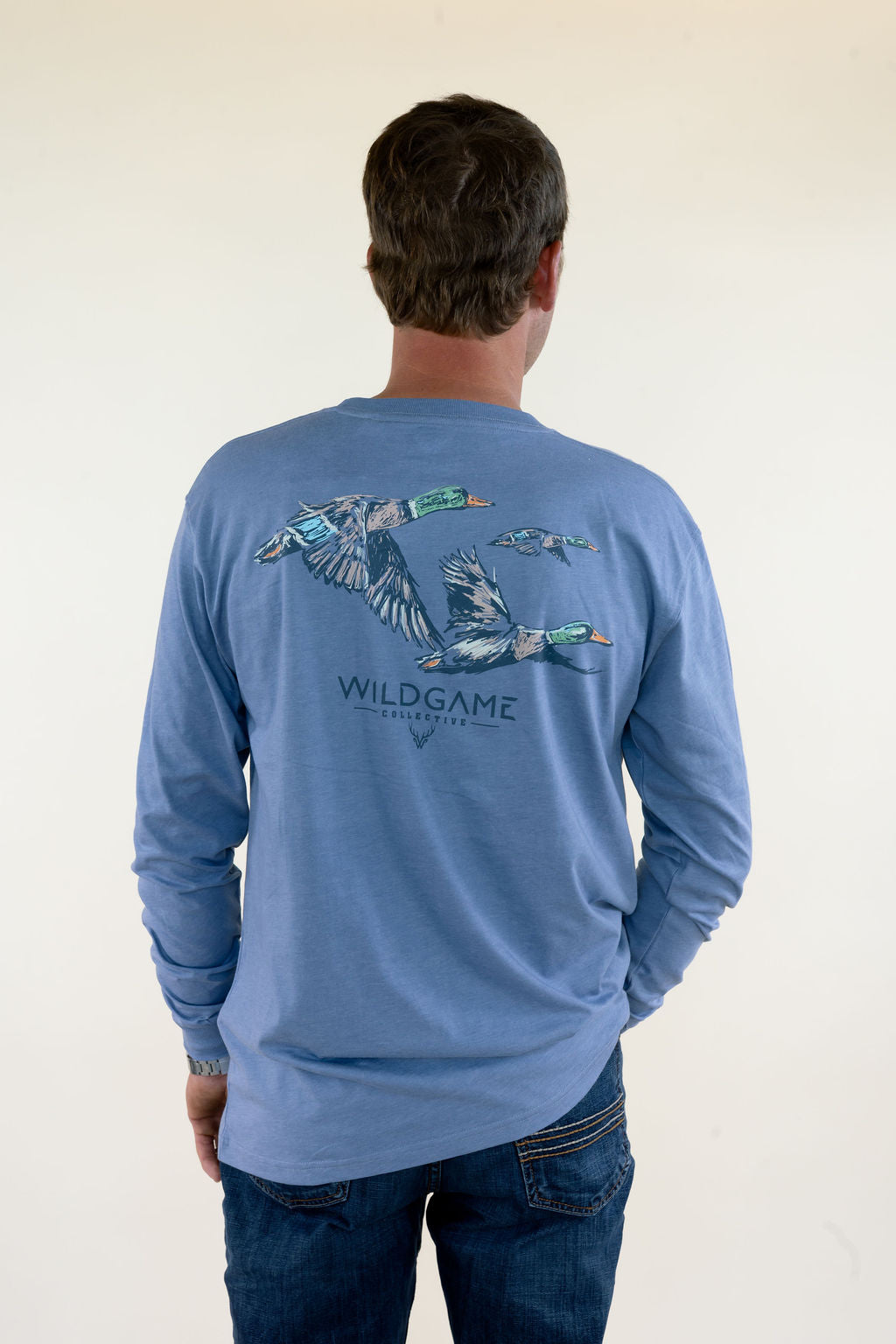 Man modeling blue Wildgame long sleeve graphic shirt featuring flying mallard ducks on the back, crew neckline, and relaxed fit