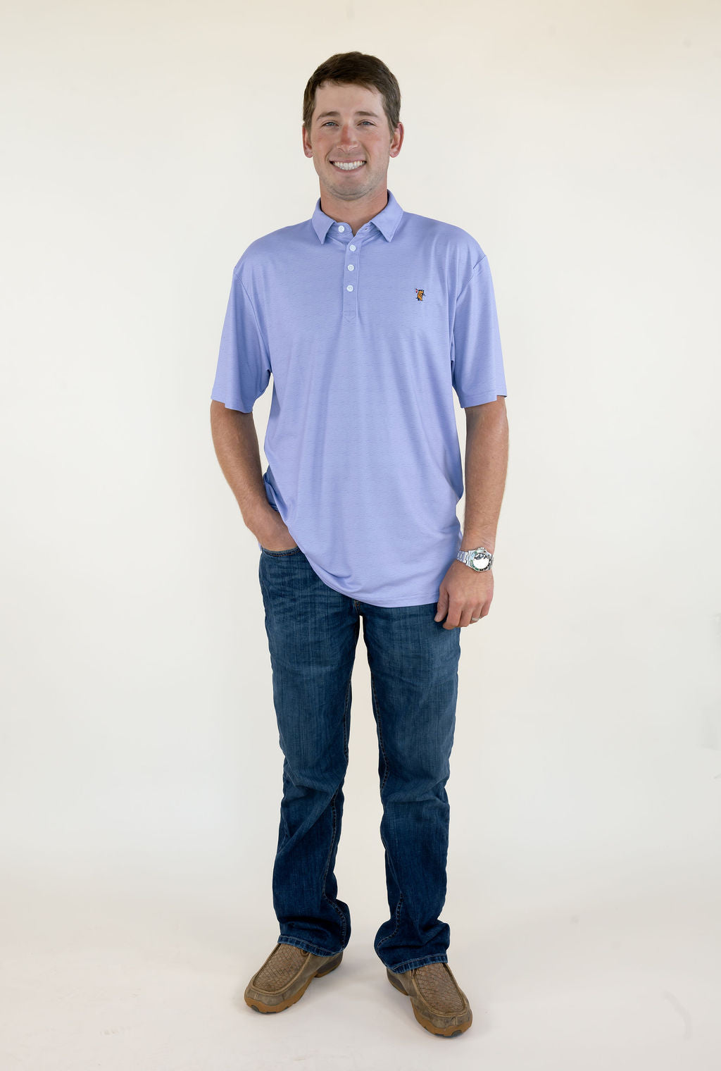 Man modeling light lavender short sleeve performance polo with collared neckline, button placket, small embroidered chest logo, blue jeans, and brown shoes
