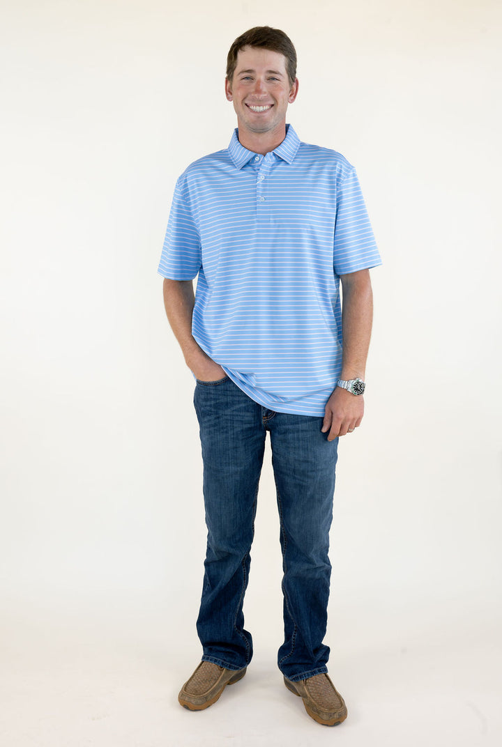 Full-length view of man wearing blue and pink striped performance polo with short sleeves, blue jeans, and brown shoes