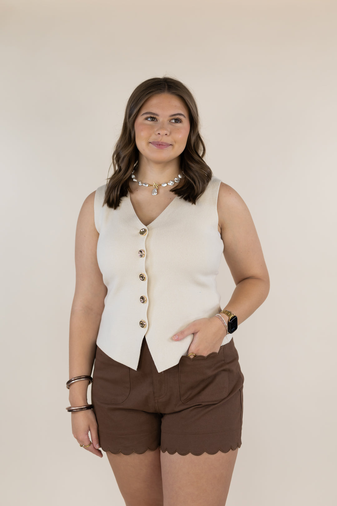 A woman modeling a sleeveless cream-colored vest with gold buttons and brown scalloped shorts against a solid beige background.