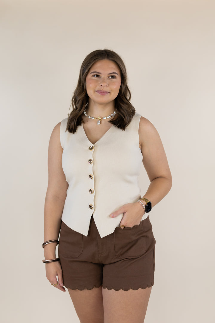 A woman modeling a sleeveless cream-colored vest with gold buttons and brown scalloped shorts against a solid beige background.