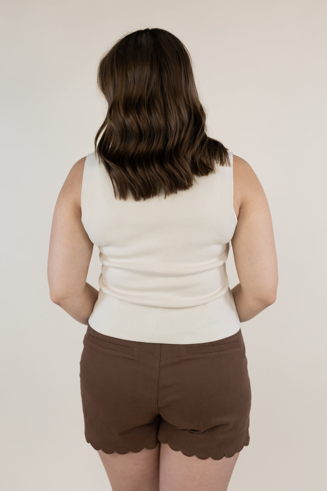 A woman shown from behind wearing a cream ribbed knit sleeveless top and brown scalloped shorts against a plain light beige background.