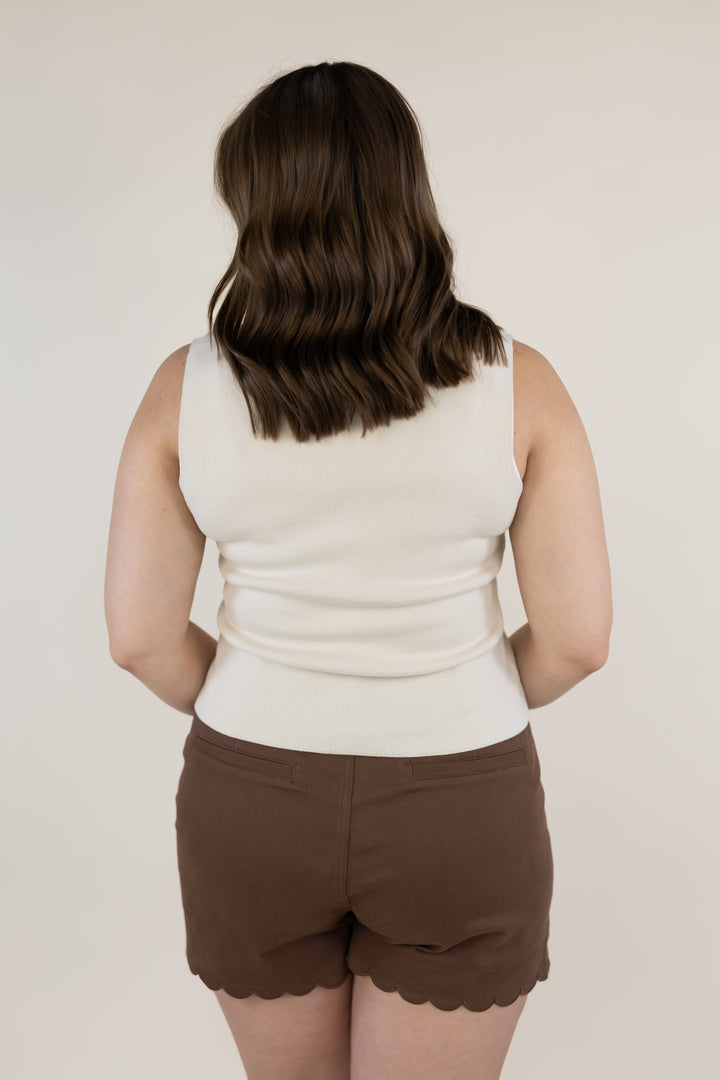A woman shown from behind wearing a cream ribbed knit sleeveless top and brown scalloped shorts against a plain light beige background.