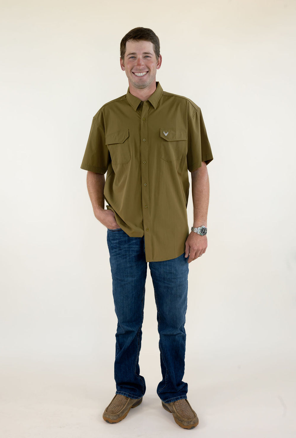 Male model wearing the WILDGAME Maverick short sleeve olive performance button down shirt with blue jeans, front full-body view.