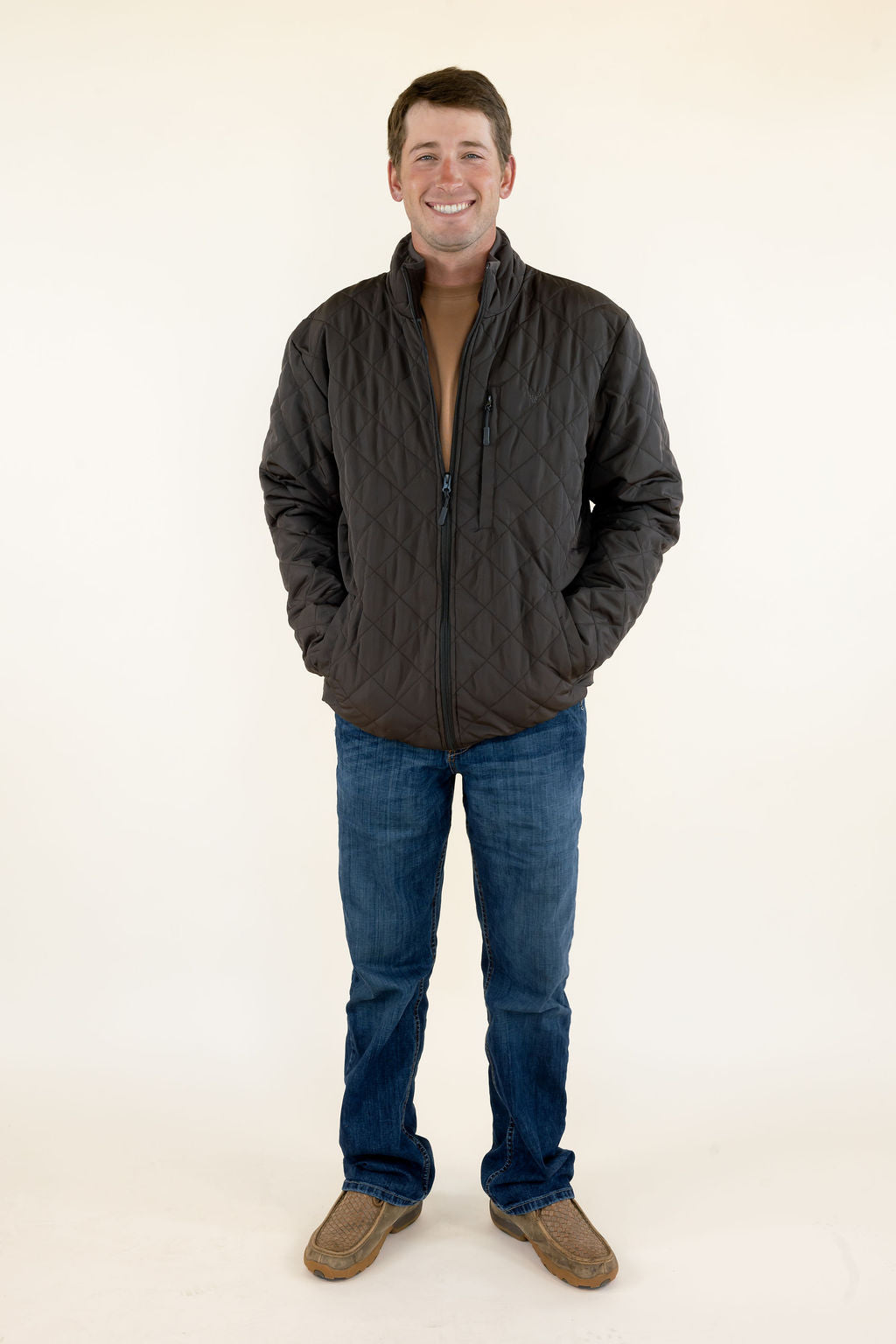 Male model wearing the WILDGAME Gambler brown quilted jacket with blue jeans, full-body front view.