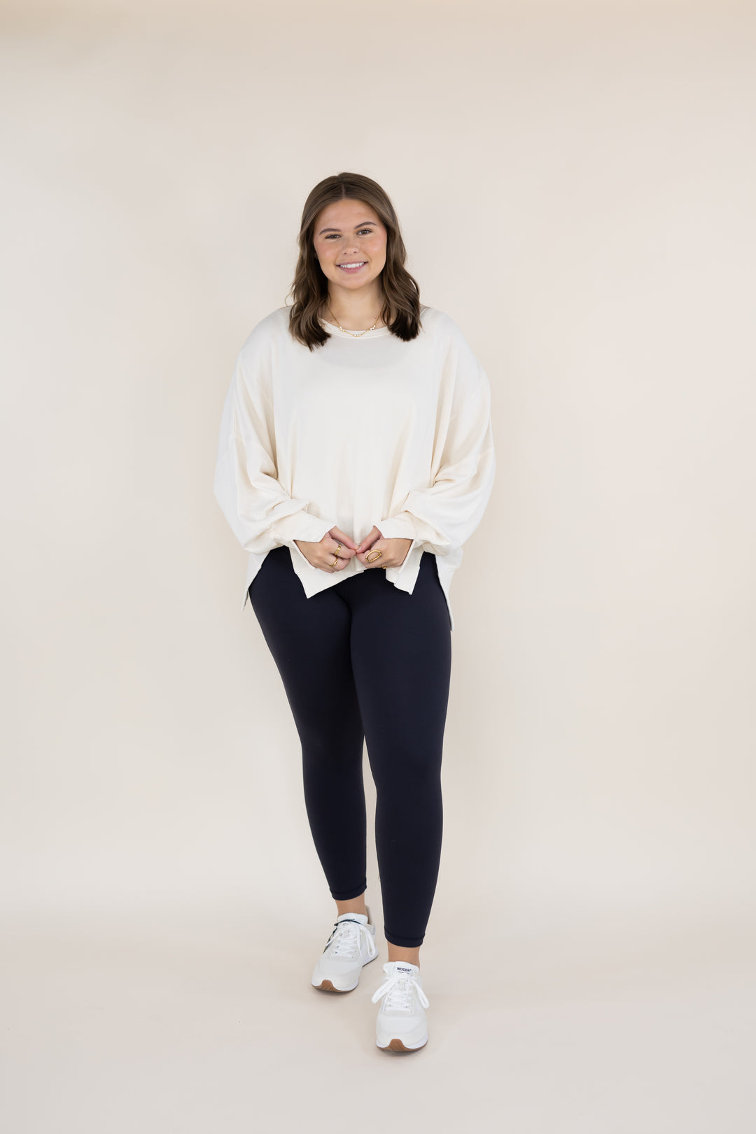 A woman modeling a cream-colored oversized long-sleeve top and dark navy leggings with white sneakers, standing against a solid light beige studio background.