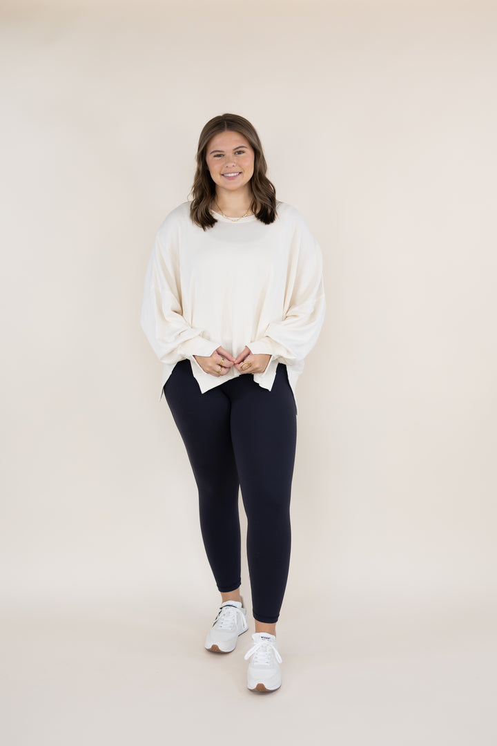 A woman modeling a cream-colored oversized long-sleeve top and dark navy leggings with white sneakers, standing against a solid light beige studio background.