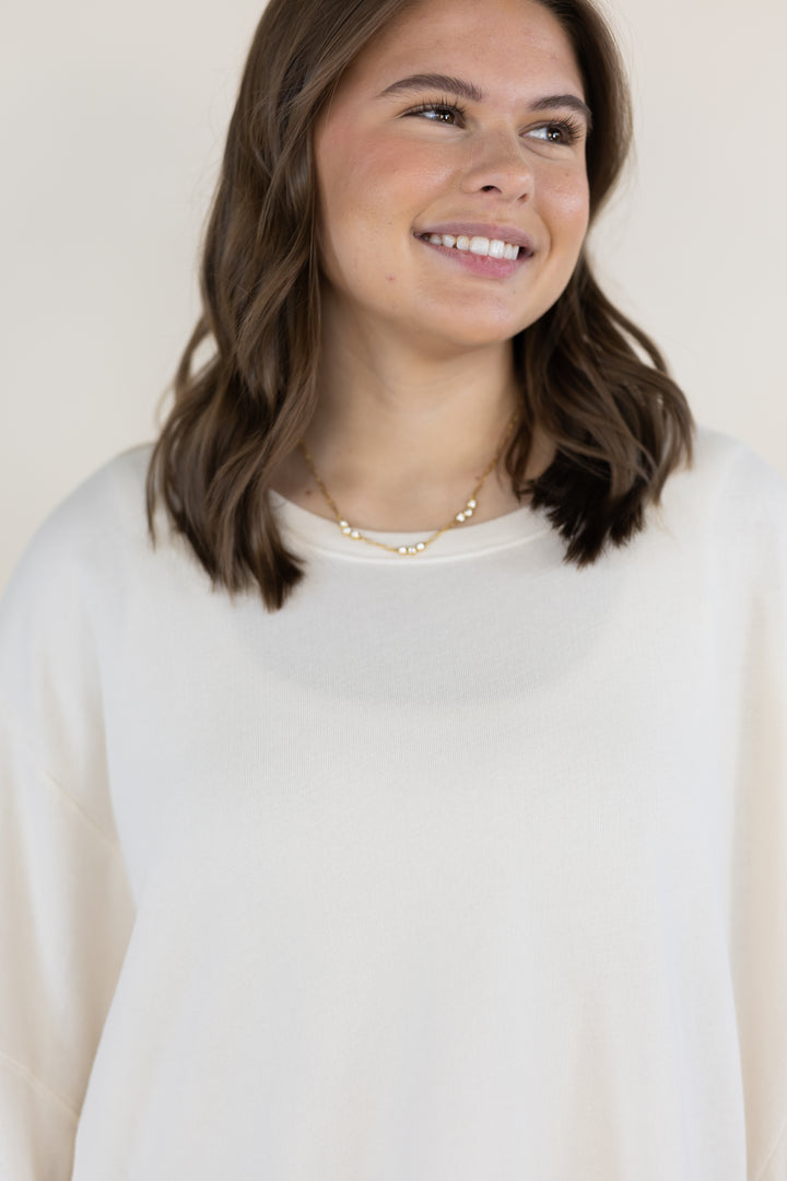 A woman with shoulder-length brown hair smiling and looking to the side, wearing a cream-colored crewneck sweater and a delicate gold necklace, set against a plain light beige background.