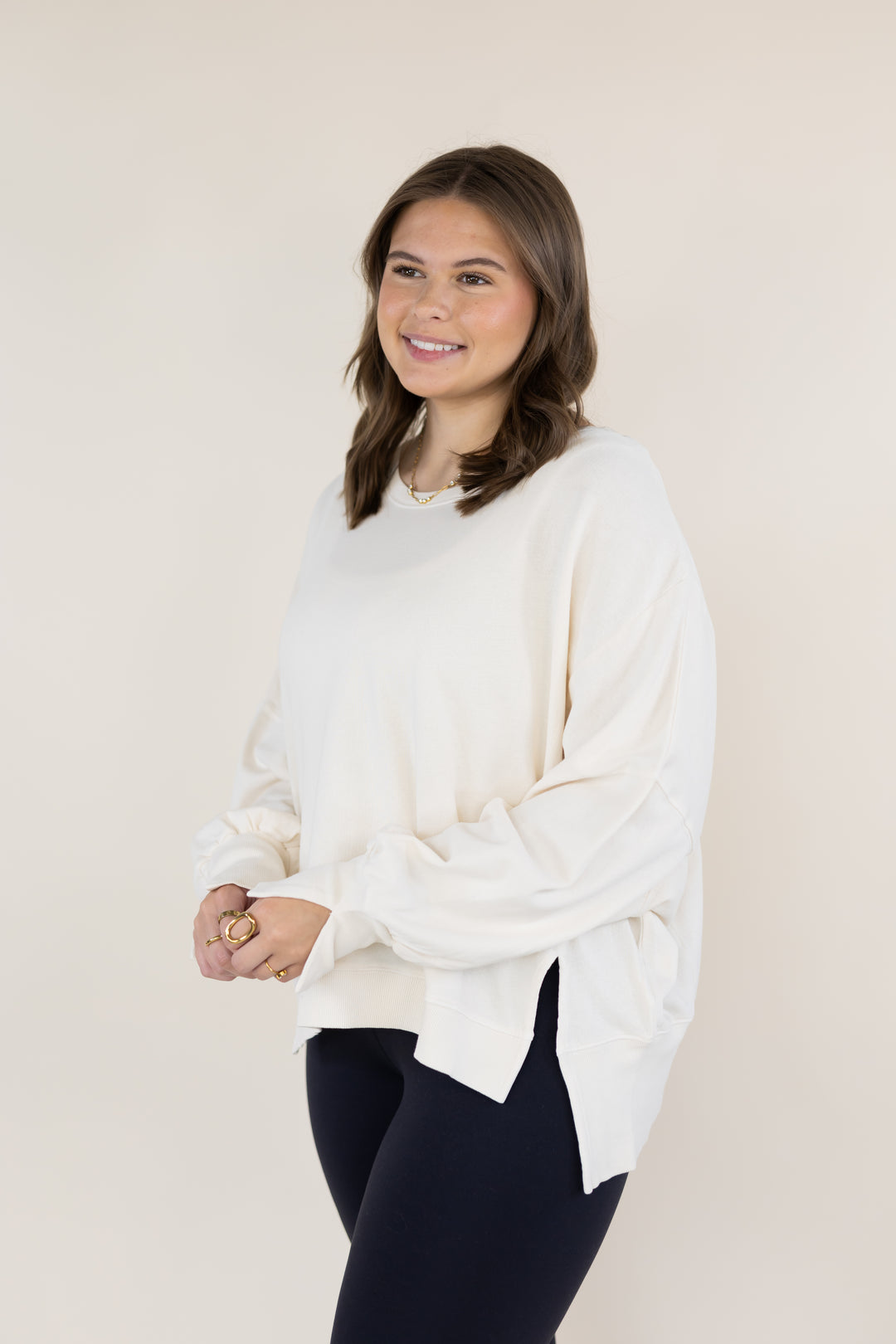A woman modeling a cream-colored, oversized long-sleeve top with a side slit, paired with dark leggings, against a solid light beige background.