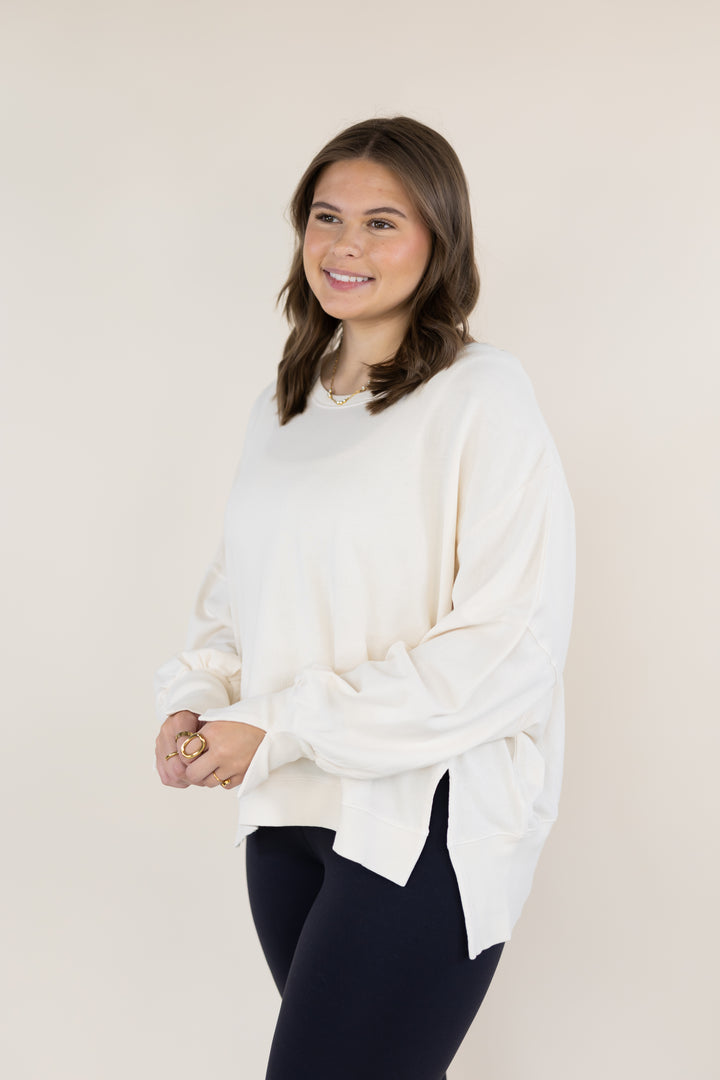 A woman modeling a cream-colored, oversized long-sleeve top with a side slit, paired with dark leggings, against a solid light beige background.