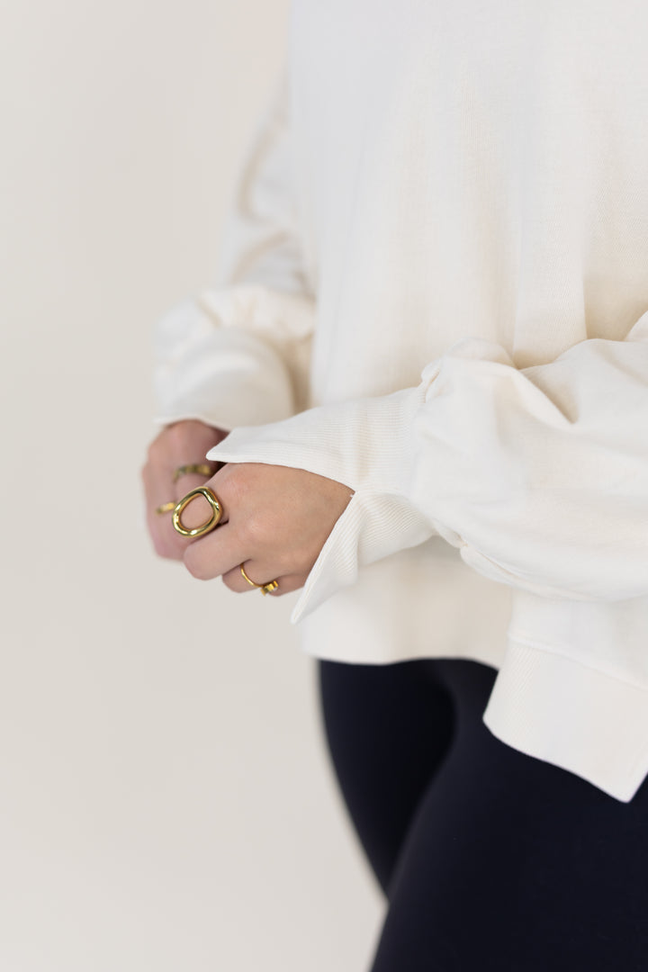 A close-up lifestyle shot of a person wearing a white long-sleeve top and black leggings, highlighting a large gold ring on their hand.