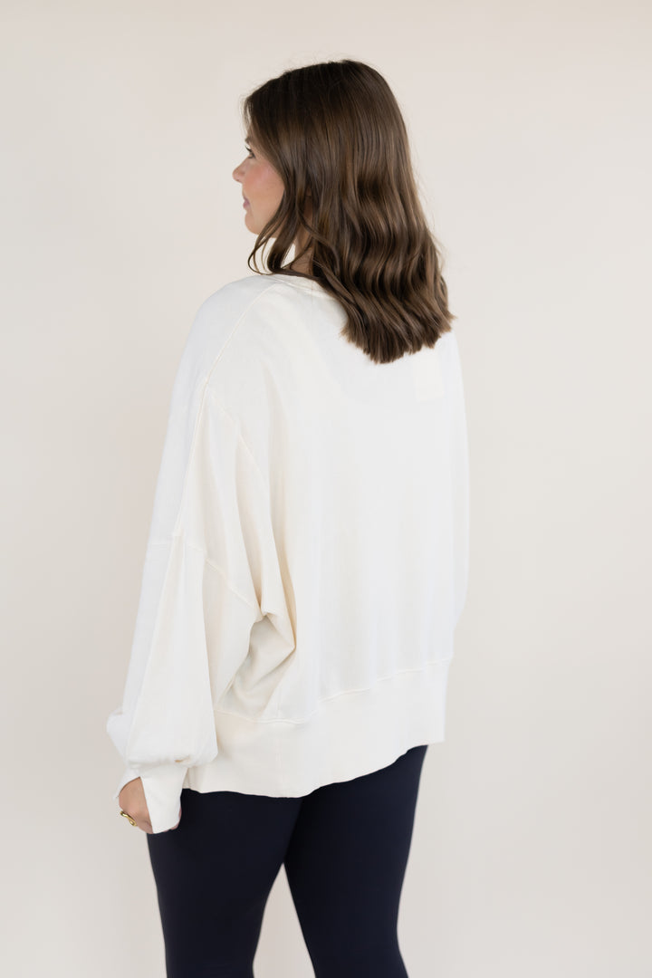 A woman with shoulder-length brown hair is shown from the back wearing a loose white long-sleeve top and black leggings, standing against a plain light beige studio background.
