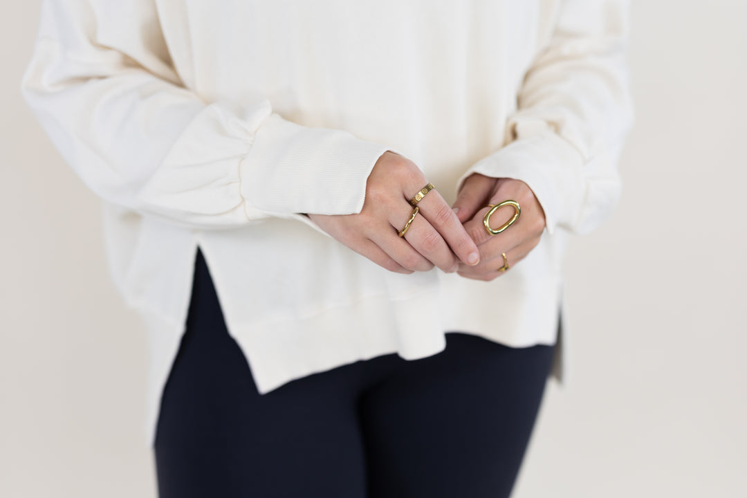 A close-up of a person wearing multiple gold rings, including a large oval ring and smaller band rings, styled with a cream-colored long-sleeve top and dark pants against a light background.