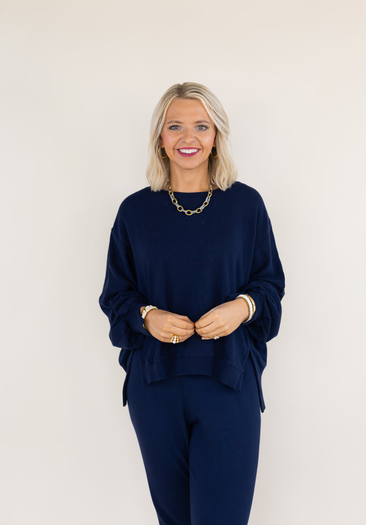 A smiling blonde woman wearing a matching navy blue lounge set, consisting of a long-sleeve top and pants, styled with a gold chain necklace and bracelets, standing against a plain off-white background.