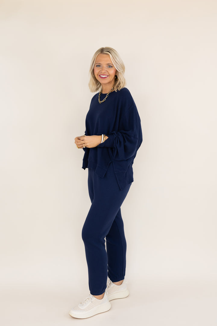 A woman modeling a matching navy blue lounge set consisting of a long-sleeve top and pants, paired with white sneakers, against a solid light beige background.