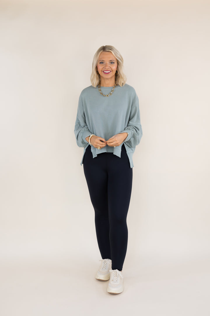 A woman modeling a sage long-sleeve knit top with dark navy leggings and white sneakers, standing against a plain off-white studio background.