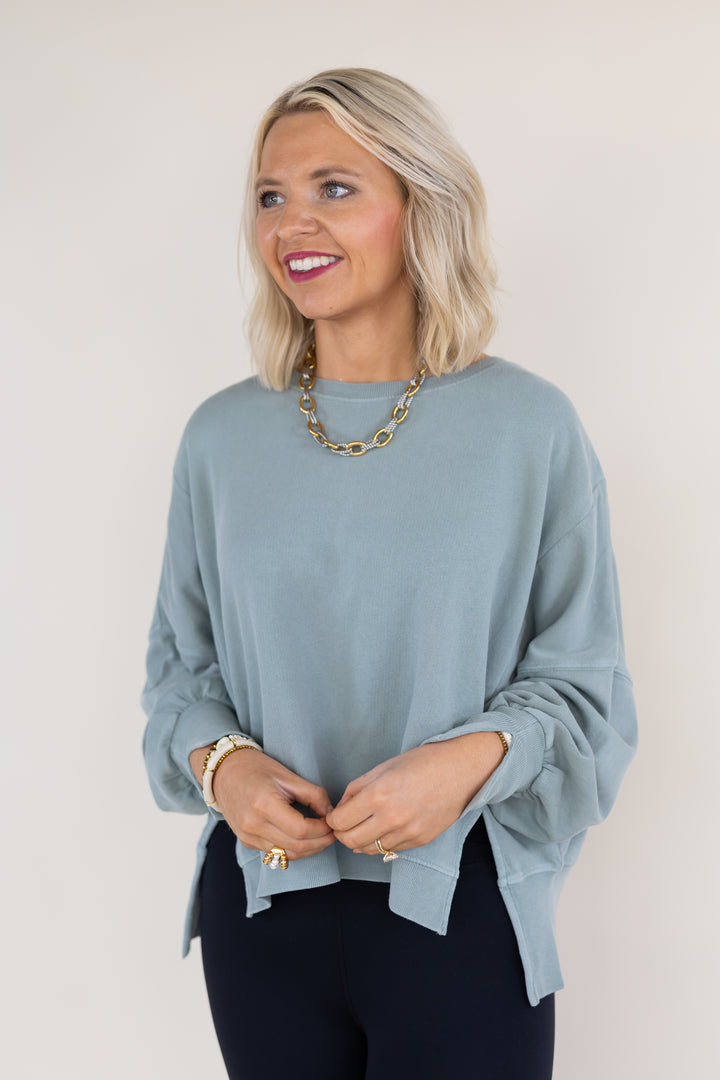 A woman with blonde hair wearing a sage long-sleeve top and dark pants, accessorized with a gold chain necklace and bracelets, posing against a plain light beige background.