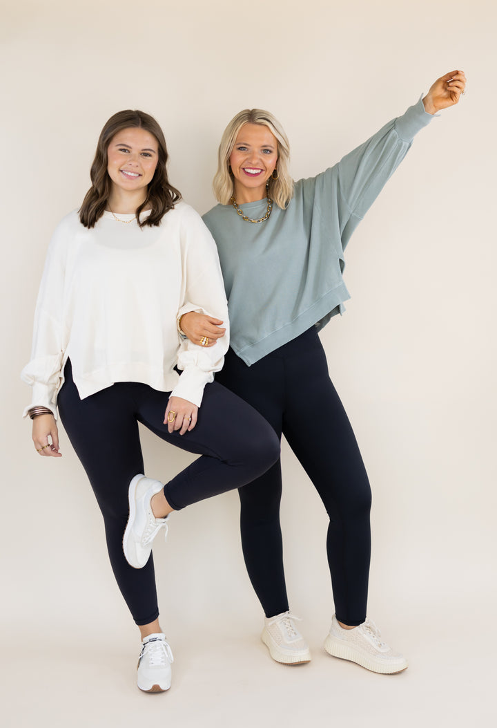 Two women modeling matching dark leggings and sweatshirts against a plain beige studio background. The woman on the left wears a white sweatshirt and white sneakers, while the woman on the right wears a sage green sweatshirt, a gold necklace, and white sneakers.
