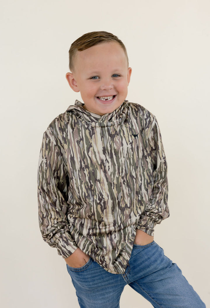 Young boy modeling a youth performance camo hoodie with long sleeves, relaxed fit, and earthy camouflage print, paired with blue jeans.