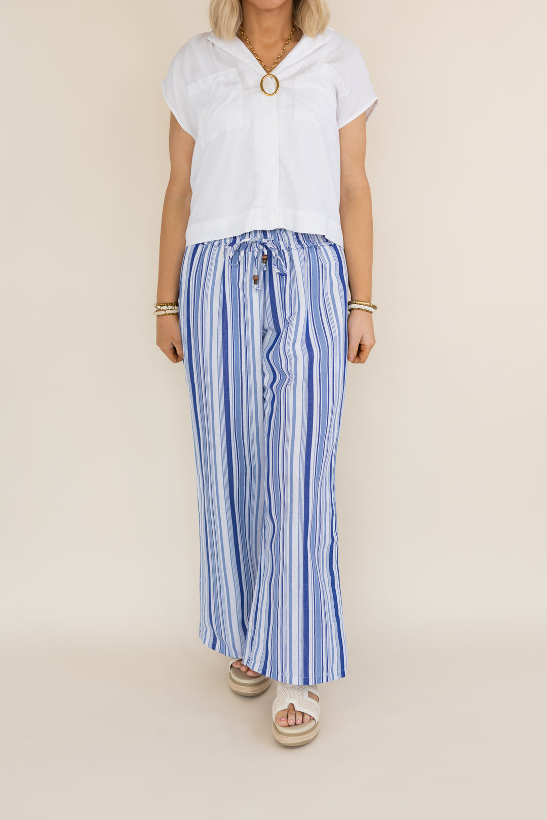 A woman modeling a white short-sleeve top with a gold ring detail at the neckline and blue-and-white vertically striped wide-leg drawstring pants, paired with beige slide sandals, against a plain beige studio background.