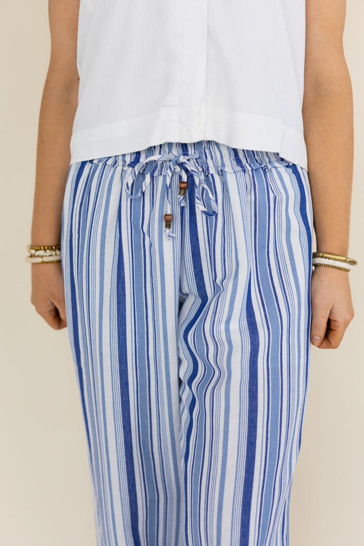 A person wearing loose blue and white vertically striped drawstring pants with a white sleeveless top, shown from the chest down against a plain light beige background.