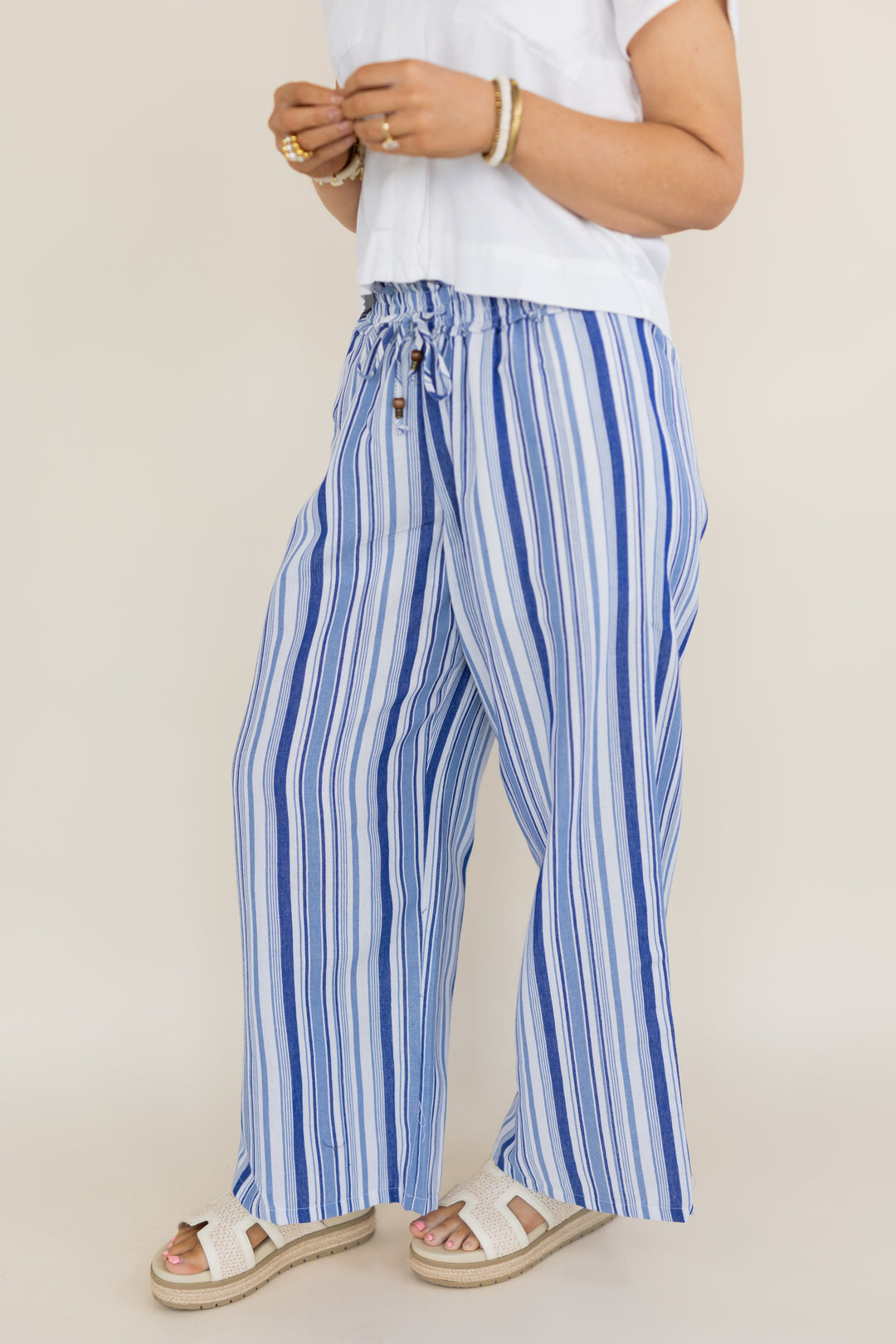 A person wearing wide-leg drawstring pants with vertical blue and white stripes, paired with a white top and white slide sandals, photographed against a plain light beige studio background.