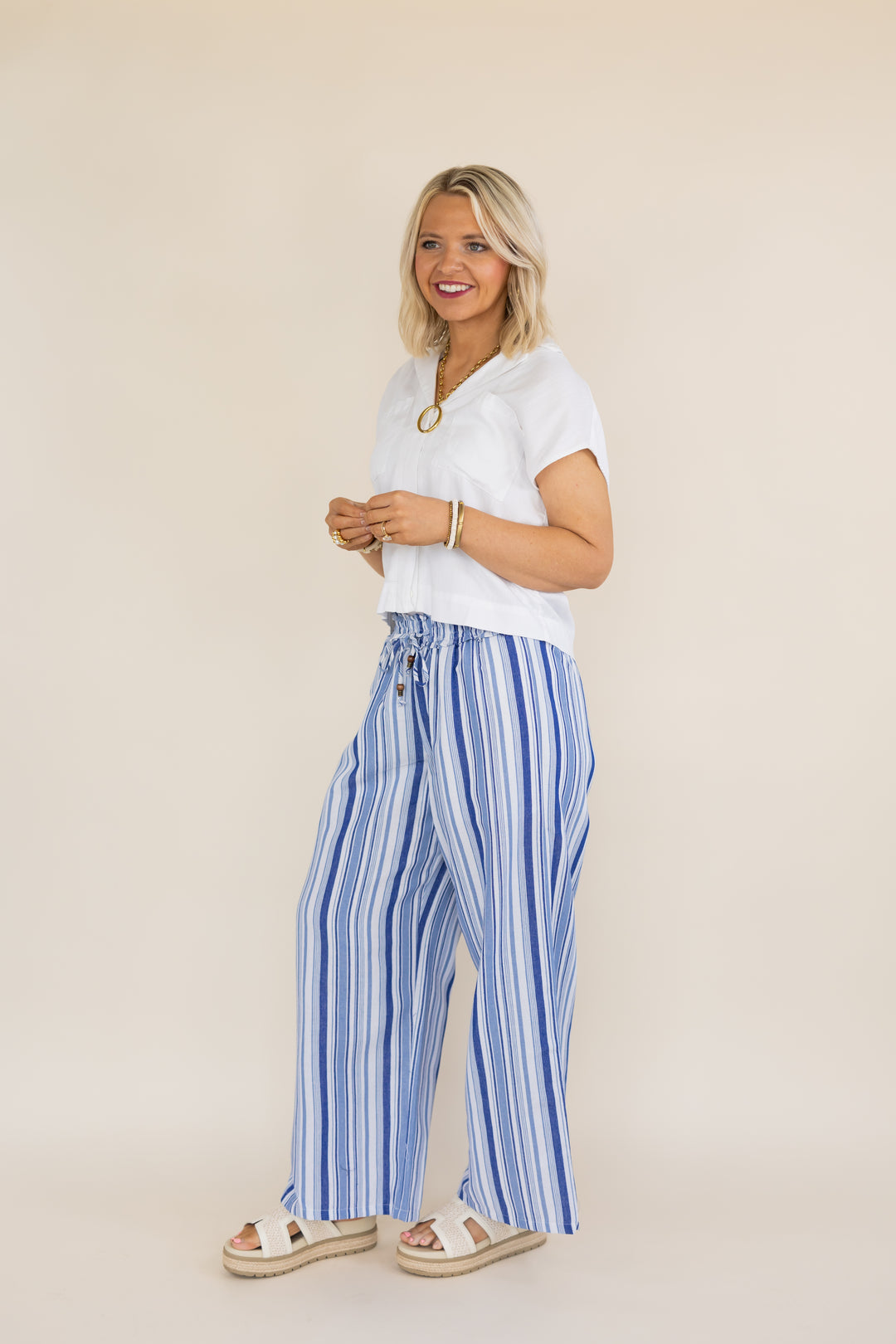 A woman modeling a white short-sleeve top and blue-and-white vertically striped wide-leg pants with beige sandals, standing against a plain beige studio background.